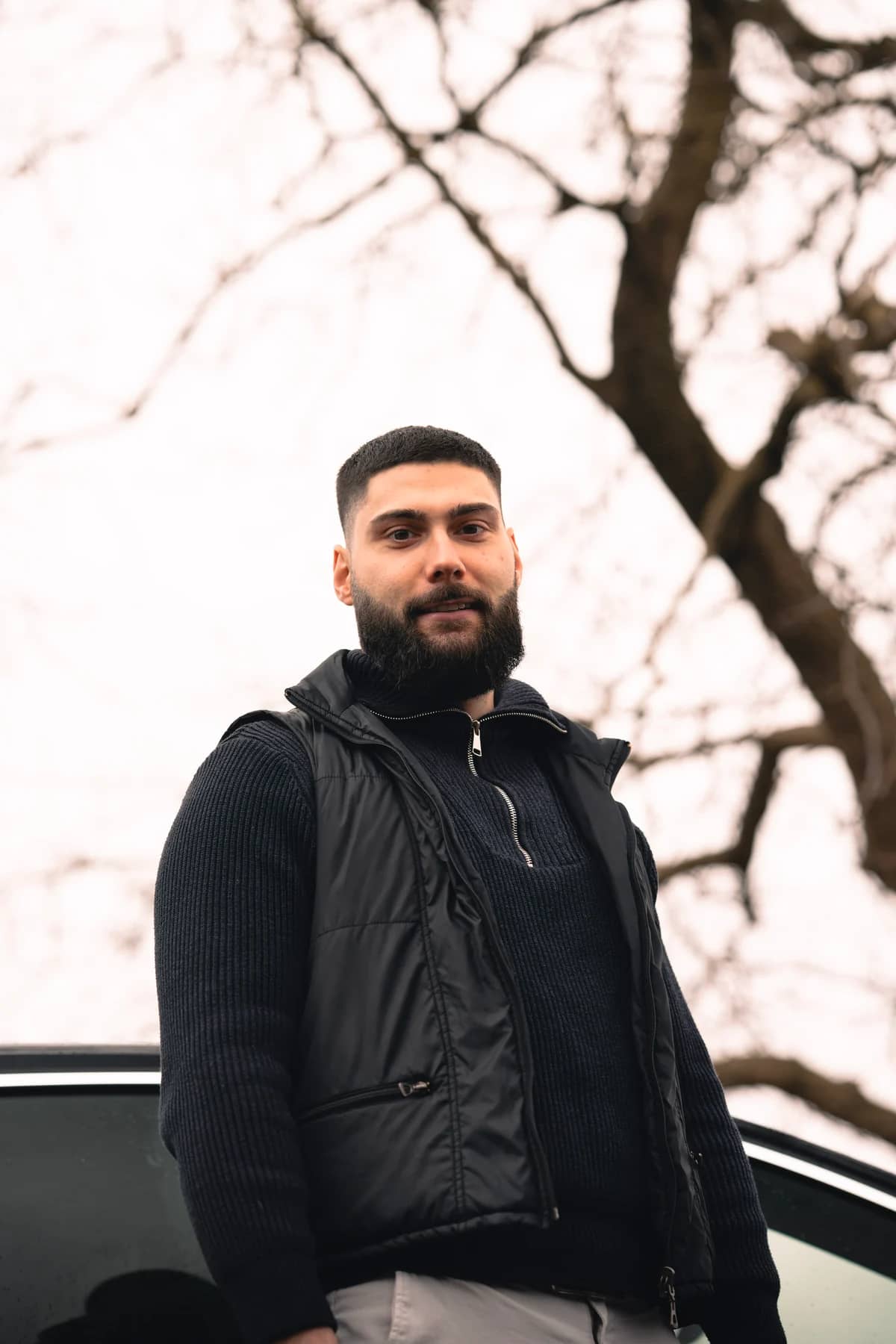 A DERLO CP contact person in a black vest stands next to a car and looks into the camera; a tree in the background.