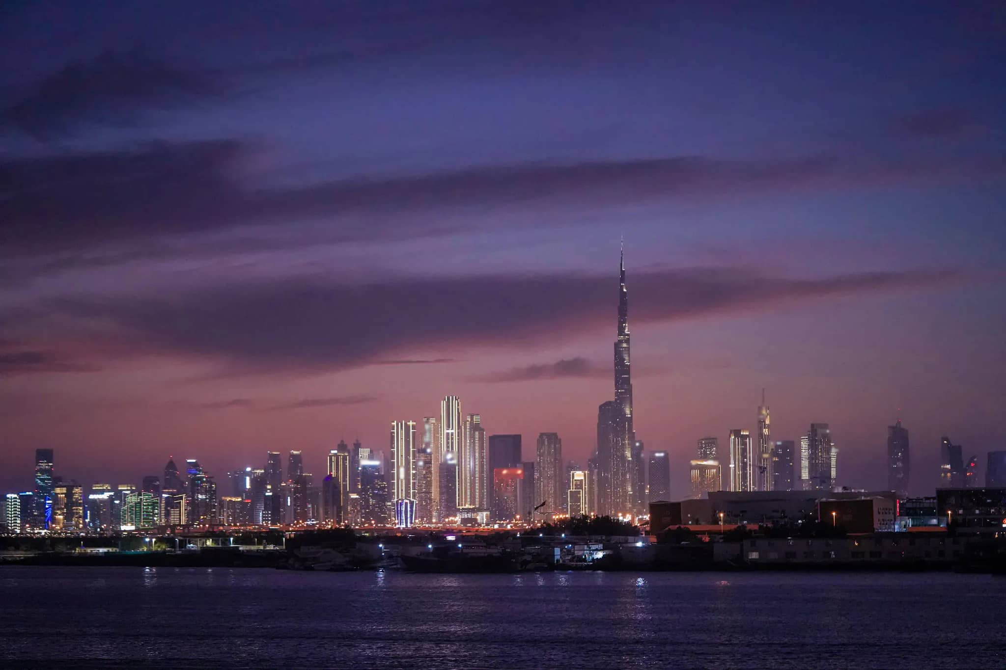 Skyline von Dubai in der Abenddämmerung unter violettem Wolkenhimmel, rechts der Mitte der Burj Khalifa, davor dunkles Wasser.