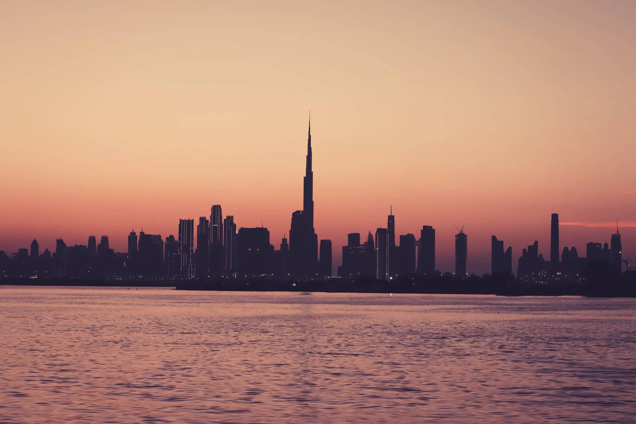 Silhouette der Skyline von Dubai mit dem Burj Khalifa vor warmem Abendhimmel nach Sonnenuntergang, im Vordergrund ruhiges Wasser.