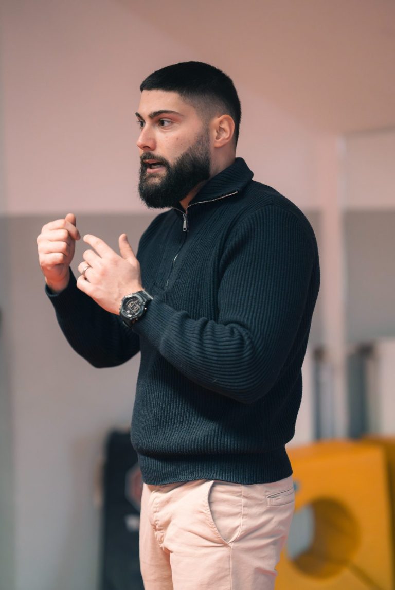 Mann mit Bart hält einen Vortrag, gekleidet in Pullover und beigefarbene Hose.