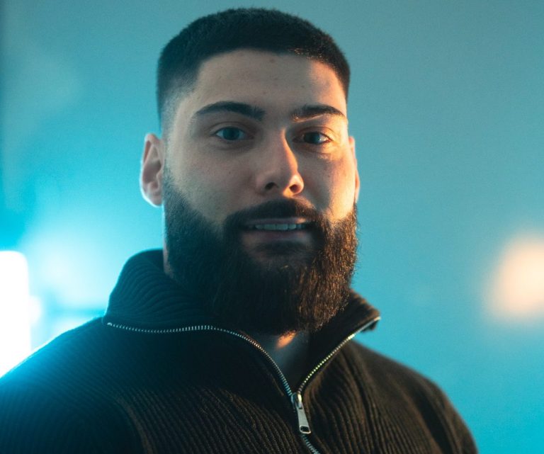 Junger Mann mit Bart, lächelnd vor einem blauen Hintergrund.