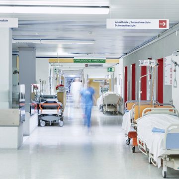 Klinikflur mit Betten, Rollstühlen und einem Arzt in Schutzkleidung.