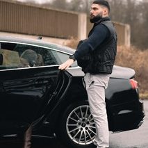 Mann in Weste öffnet die Tür eines schwarzen Autos in einer grauen Umgebung.