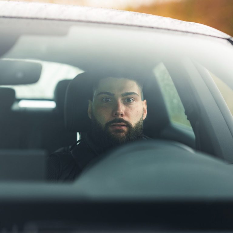 Mann mit Bart sitzt am Steuer eines Autos und schaut in den Rückspiegel.