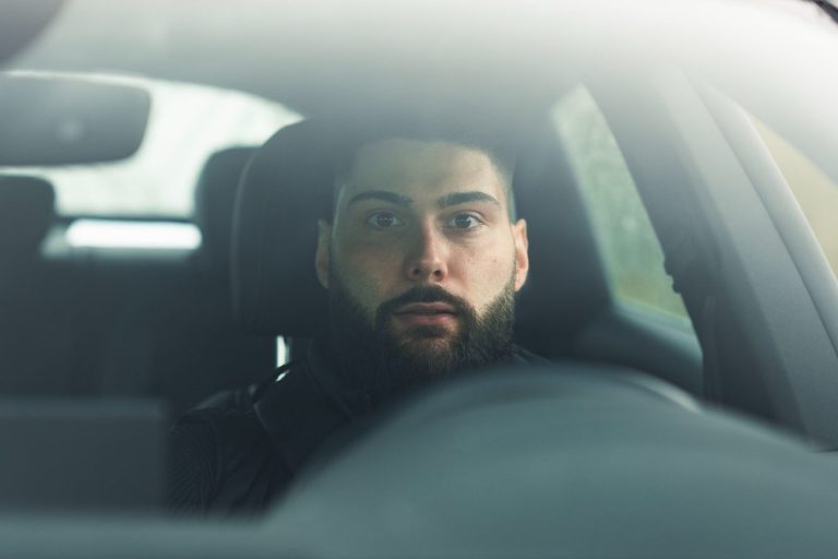 Junger Mann mit Bart sitzt am Steuer eines Autos und schaut überrascht.