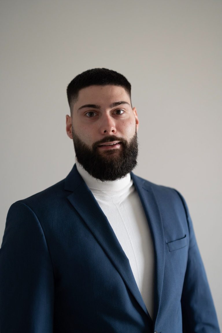 Mann mit Bart in blauer Jacke und weißem Rollkragenpullover vor neutralem Hintergrund.