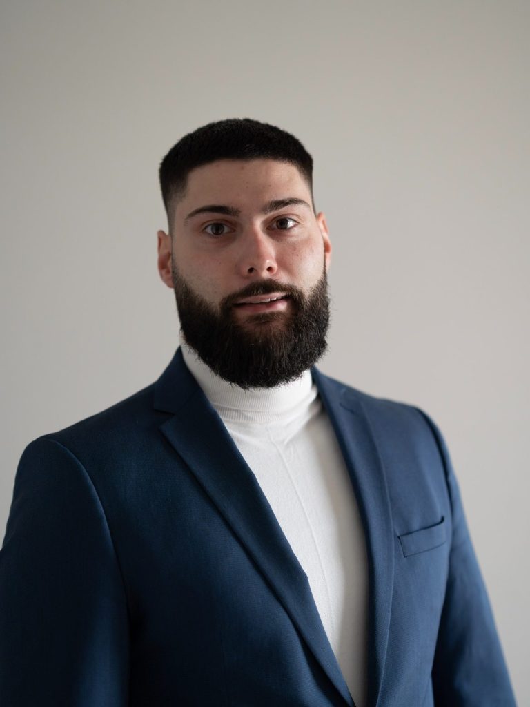 Mann mit Bart in blauer Jacke und weißen Rollkragenpullover vor neutralem Hintergrund.