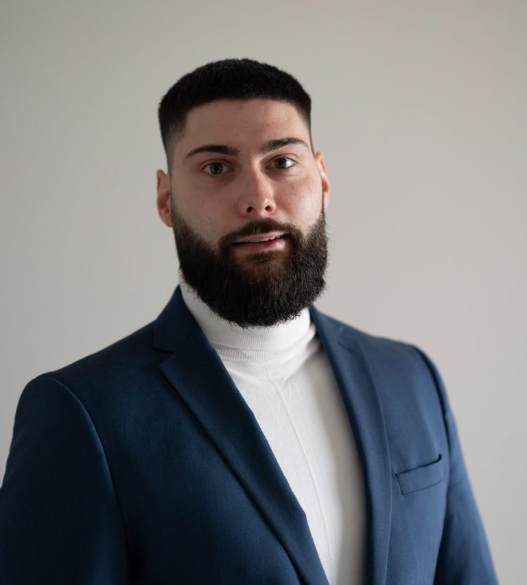 Mann mit kurzem Bart und dunklem Anzug steht vor neutralem Hintergrund.