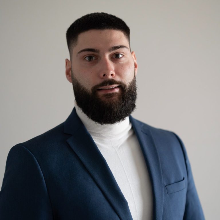 Mann mit Bart trägt einen blauen Anzug und weißen Rollkragenpullover.