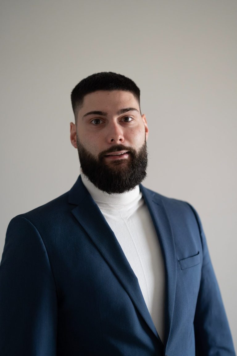 Mann mit Bart in einem blauen Anzug und hellem Rollkragenpullover vor grauem Hintergrund.