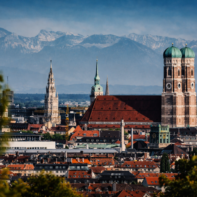 Panoramablick auf München mit den Alpen im Hintergrund und historischen Gebäuden.