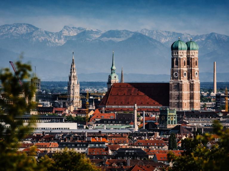 Panoramablick auf München mit den Alpen im Hintergrund und markanten Kirchenkuppeln.