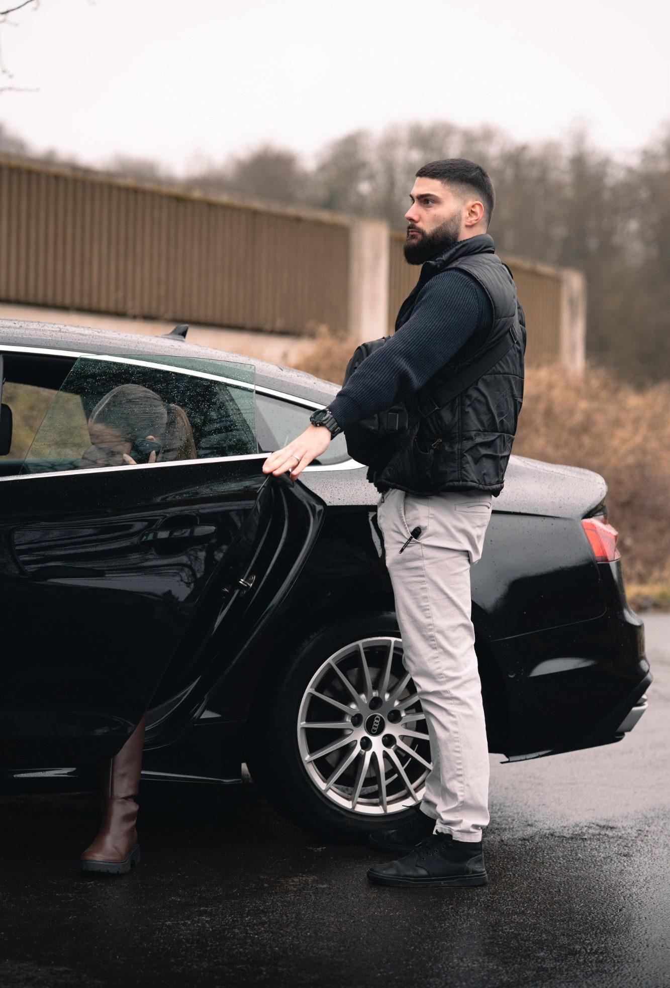 Mann steht neben einem Auto und öffnet die hintere Tür, während es regnet.