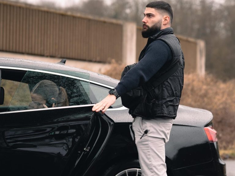 Mann in dunkler Weste steht neben einem schwarzen Auto, schaut zur Seite. Hund im Inneren.