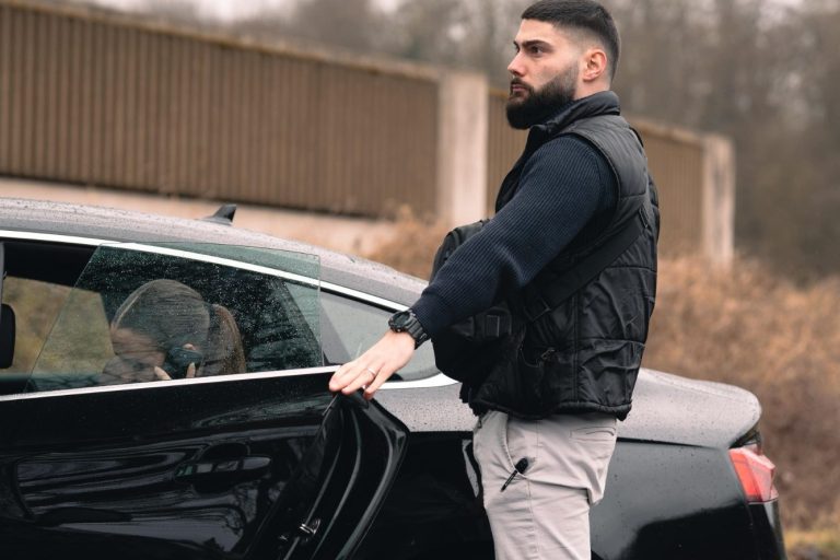 Ein Mann mit Bart öffnet die Tür eines schwarzen Autos in einer ländlichen Umgebung.